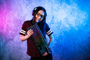 Beautiful Friendly Pro Gamer Streamer Girl Posing With a Keyboard in Her Hands, Wearing Glasses. Attractive Geek Girl with Cool Neon Retro Colors in Background. - 435412387