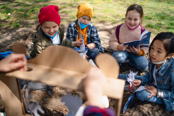 Teacher with small children sitting outdoors in city park, learning group education and coronavirus concept.