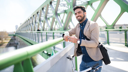 Young business man commuter with bicycle going to work outdoors in city, using smartphone on bridge.