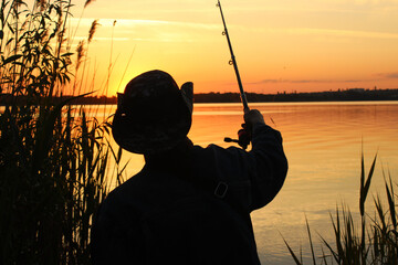 A young male fisherman in a hat throws a fishing rod into the river at sunset. Silhouette of a man...