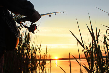 Close-up of a fishing rod with a reel in male hands against the backdrop of the sunset on the river...