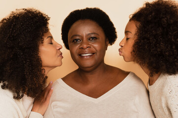 Obraz premium african american daughter and granddaughter kissing grandmother with closed eyes on beige background