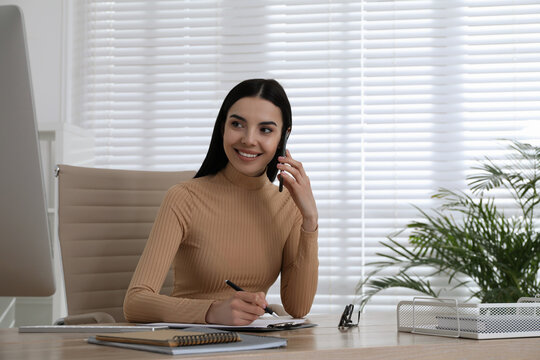 Secretary talking on smartphone at wooden table in office