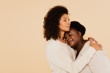 african american adult daughter hugging middle aged mother with closed eyes isolated on beige