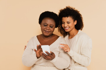 african american adult daughter pointing with finger to smartphone in hands of middle aged mother on beige background