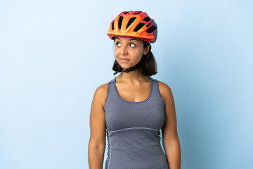 Young cyclist woman isolated on blue background making doubts gesture looking side - obrazy, fototapety, plakaty