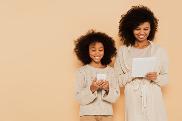 smiling african american preteen daughter and adult mother holding cellphone and tablet in hands isolated on beige