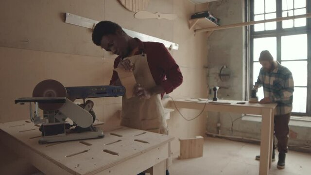 Medium shot of African-American male carpenter standing at workbench and sanding board edges using grinding machine while his male Caucasian colleague working on background