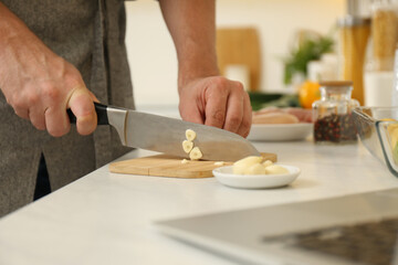 Obraz na płótnie Canvas Man cutting garlic while watching online cooking course via laptop in kitchen, closeup