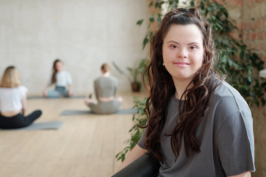 Brunette female with Down syndrome looking at you while group of girls practicing yoga in gym
