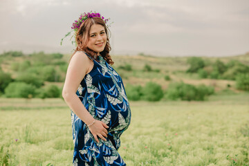 Pregnancy and maternity. Young pregnant woman in wreath of flowers standing in field, holding her big belly and posing for camera