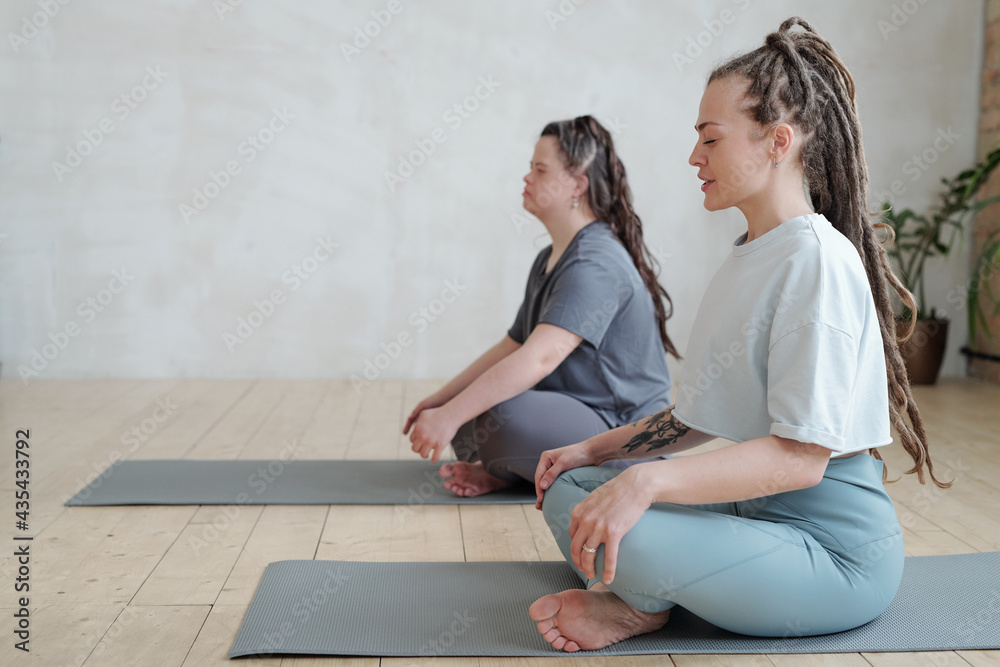 Wall mural Trainer and disable girl with Down syndrome sitting on mats with their legs crossed during yoga exercise