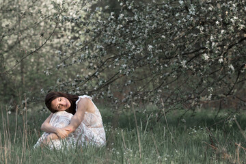 a beautiful young girl in a white dress sits with her eyes closed under a blooming apple tree