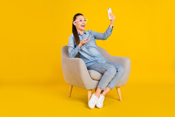 Photo of impressed cute lady dressed jeans shirt spectacles sitting chair talking modern device smiling isolated yellow color background