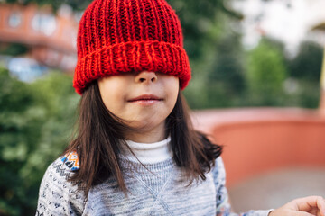 Portrait of a cute little girl in a red knitted hat that covers her eyes during resting outside after school. A pretty kid have fun outdoor.