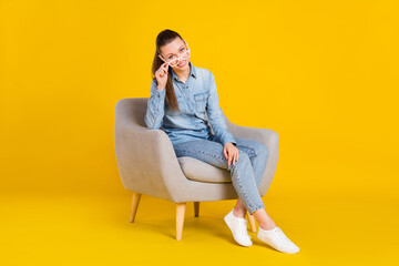 Fototapeta na wymiar Photo of cute adorable young woman wear denim shirt arm glasses sitting chair isolated yellow color background