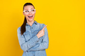 Photo of cute impressed young lady dressed jeans shirt spectacles pointing finger empty space smiling isolated yellow color background