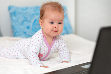 Fototapeta premium Happy baby in sliders is lying on the bed and watching cartoons on a laptop. The concept of entertainment and education for children using the Internet