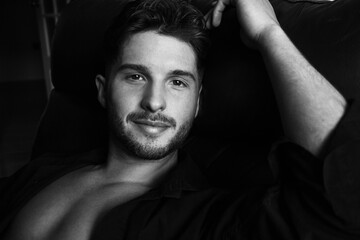Obraz premium Portrait of attractive young man with beard, open shirt revealing chest, sitting in leather armchair, looking at camera
