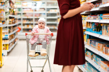 Shopping. A toddler sits in a grocery cart and looks around while his mother picks out groceries. Family shopping in the supermarket