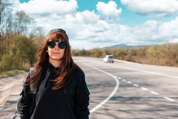 Portrait of caucasian young woman with backpack and sunglasses on the empty road. Copy space. The concept of local traveling and hitchhiking