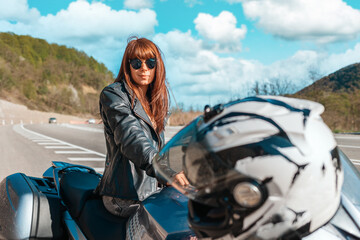 World Motorcyclist Day. A young caucasian woman in a leather jacket and sunglasses sitting on a motorbike. Road and cloudscape on the background. Motorcycle local travel concept