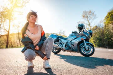 Fototapeta na wymiar Pretty young woman with tattoos on her arm, wearing leather jacket, and posing sitting. Road and motorbike in the background. Motorcycle local travel concept