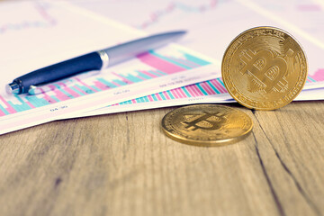 One gold coin of the bitcoin cryptocurrency stands on its own end, the second one lies on the charts of changes in cryptocurrency rates on a wooden table in the office