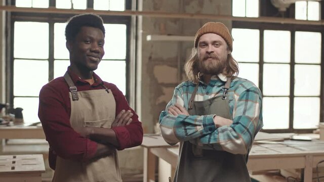 Medium shot portrait of cheerful African and Caucasian joiners wearing aprons standing in carpentry workshop, looking at camera and smiling