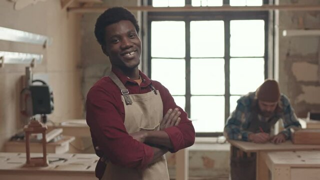 Medium shot portrait of cheerful young African joiner standing in carpentry workshop and looking at camera while his Caucasian coworker working on background
