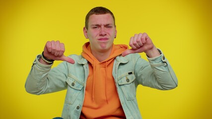 Upset handsome stylish teen man in denim jacket showing thumbs down sign gesture, expressing discontent, disapproval, dissatisfied, dislike. Young adult guy. Indoor studio shot on yellow background