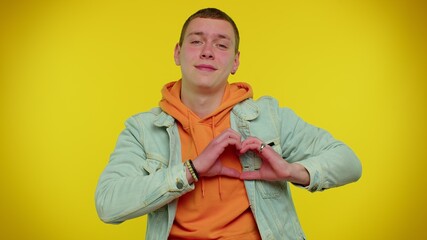 Smiling cute teenager student boy in denim jacket makes heart gesture demonstrates love sign expresses good feelings and sympathy. Young fashionable man isolated on yellow studio background