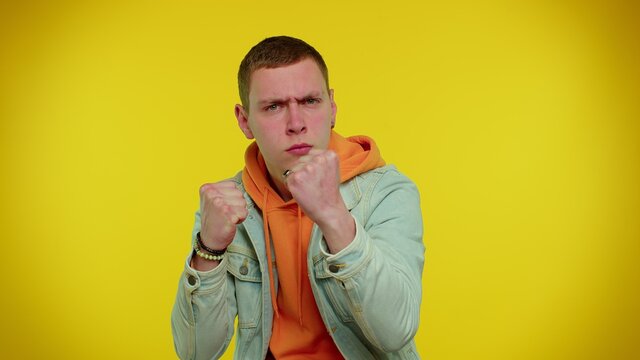 Aggressive Angry Sincere Teen Student Boy 20s Years Old In Denim Jacket Trying To Fight At Camera, Shaking Fist, Boxing With Expression, Punishment. Young Adult Man Isolated On Yellow Background