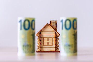 A wooden house on the background of blurred paper banknotes with a face value of 100 euros. The...