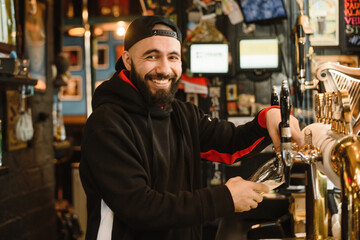 Fototapeta na wymiar smiling barman pouring beer in a bar. Bearded courageous man pours you a foaming drink. Golden tap for draft beer