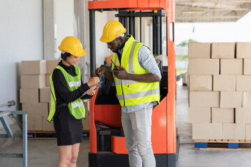 Young Asian woman with black man worker in safety vest and yellow helmet looking on document together while working at shipment at warehouse factory