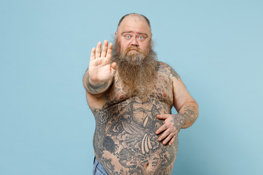 Young Serious Sad Fat Pudge Obese Chubby Overweight Blue-eyed Bearded Man Has Big Belly With Naked Tattooed Torso Showing Stop Gesture With Palm Isolated On Pastel Blue Background Studio Portrait