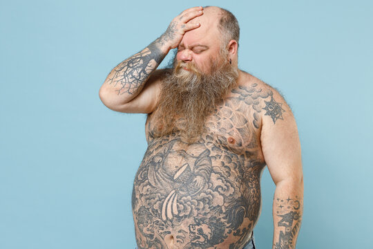 Side view sad young fat pudge obese chubby overweight blue-eyed bearded man 30s has big belly with naked tattooed torso put hand on face facepalm epic fail gesture isolated on pastel blue background.