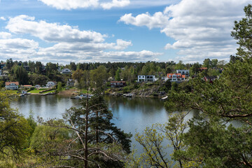 Fototapeta na wymiar Panoramic view of the Baltic Sea Bay on sunny spring day. Rocky shores of Scandinavia covered with evergreen forests. Traditional Swedish red wooden houses cabins on the coast. Blue sky white clouds.