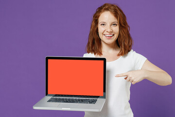 Naklejka premium Young smiling fun redhead woman 20s in white basic casual t-shirt point index finger on laptop pc computer with blank screen workspace area isolated on dark violet color background studio portrait