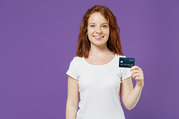 Young smiling rich successful fun happy satisfied redhead woman 20s in white basic blank print design t-shirt holding in hand credit bank card isolated on dark violet color background studio portrait