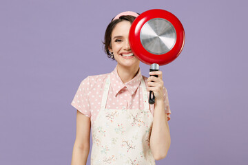Young smiling happy cheerful housewife housekeeper chef cook baker woman in pink apron cover eye with red frying pan isolated on pastel violet background studio portrait Cooking food process concept - obrazy, fototapety, plakaty