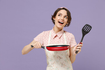 Young happy pensive housewife housekeeper chef cook baker woman in pink apron hold show red frying pan spatula look aside overhead isolated on pastel violet background Cooking food process concept - obrazy, fototapety, plakaty
