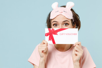 Young excited woman wear pajamas jam sleep eye mask rest relaxing at home hiding cover face with gift voucher flyer mock up isolated on pastel blue background studio. Good mood night bedtime concept.