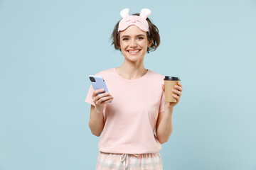 Young woman in pajamas jam sleep eye mask rest relaxing at home hold takeaway hold cup of coffee drink tea mobile cell phone isolated on pastel blue background studio. Good mood night bedtime concept.