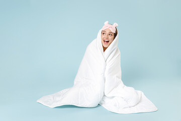 Young excited amazed woman in pajamas jam sleep eye mask rest relaxing at home sitting wrap covered under blanket duvet look aside isolated on pastel blue background. Good mood night bedtime concept.