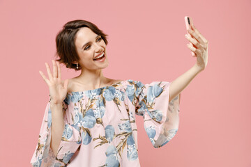 Young friendly happy woman with short hairdo wear stylish blouse doing selfie shot on mobile phone post photo on social network waving hand say hello isolated on pastel pink color background studio