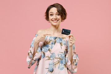 Young happy fashionable caucasian woman 20s with short hairdo wear trendy stylish blouse point index finger on credit bank card look camera isolated on pastel pink color background studio portrait