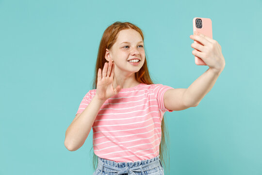 Little redhead kid girl 12-13 years old wearing pink striped tshirt do selfie shot on mobile phone post photo social network waving hand isolated on pastel blue background Lifestyle childhood concept