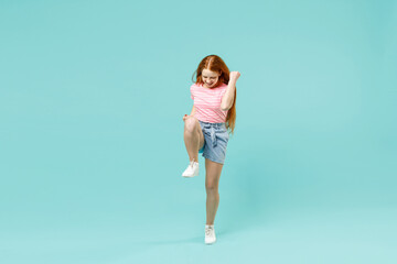 Full length little overjoyed fun redhead kid girl 12-13 years old in pink t-shirt do winner gesture clench fist raised up leg isolated on pastel blue background. Children lifestyle childhood concept.